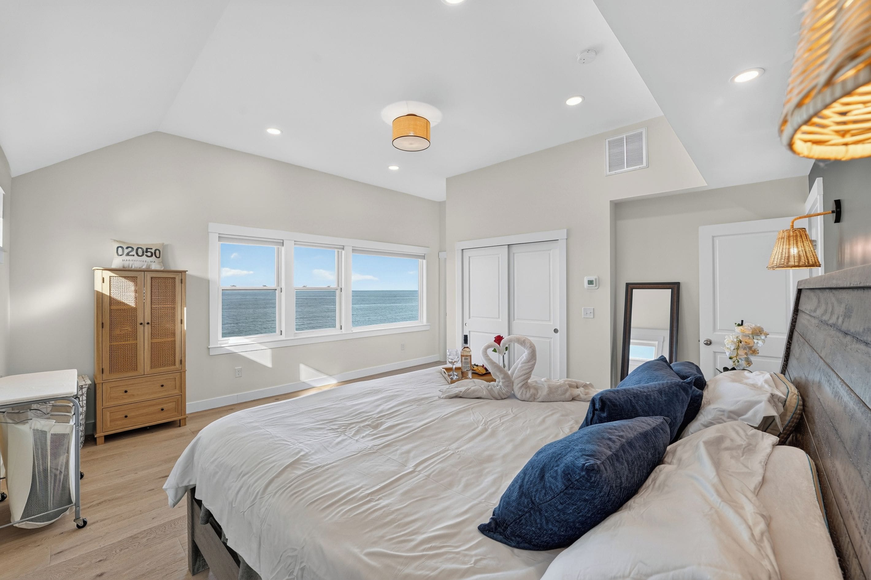 A bedroom with a large bed, a wooden wardrobe, a window with a view of the sea, and a ceiling light.