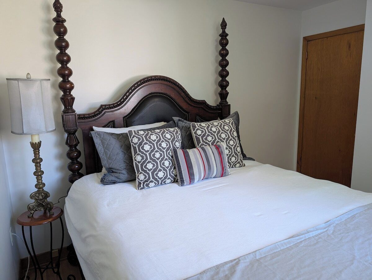Una cama con cabecera de madera, varias almohadas, una lámpara de noche y una puerta de madera.