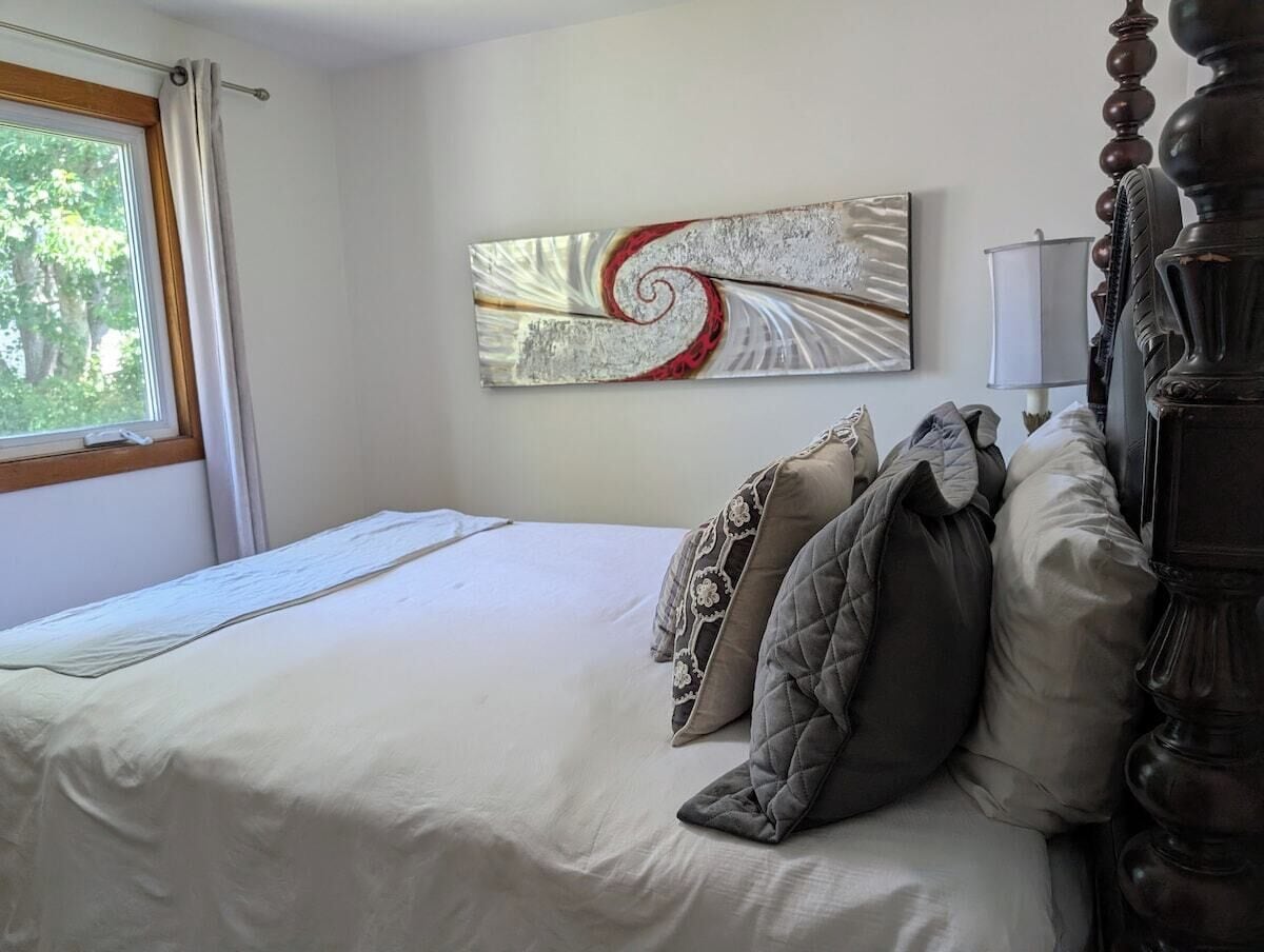 A neatly made bed with a white duvet and several pillows, a modern abstract painting on the wall, a window with curtains, and a classic wooden headboard.