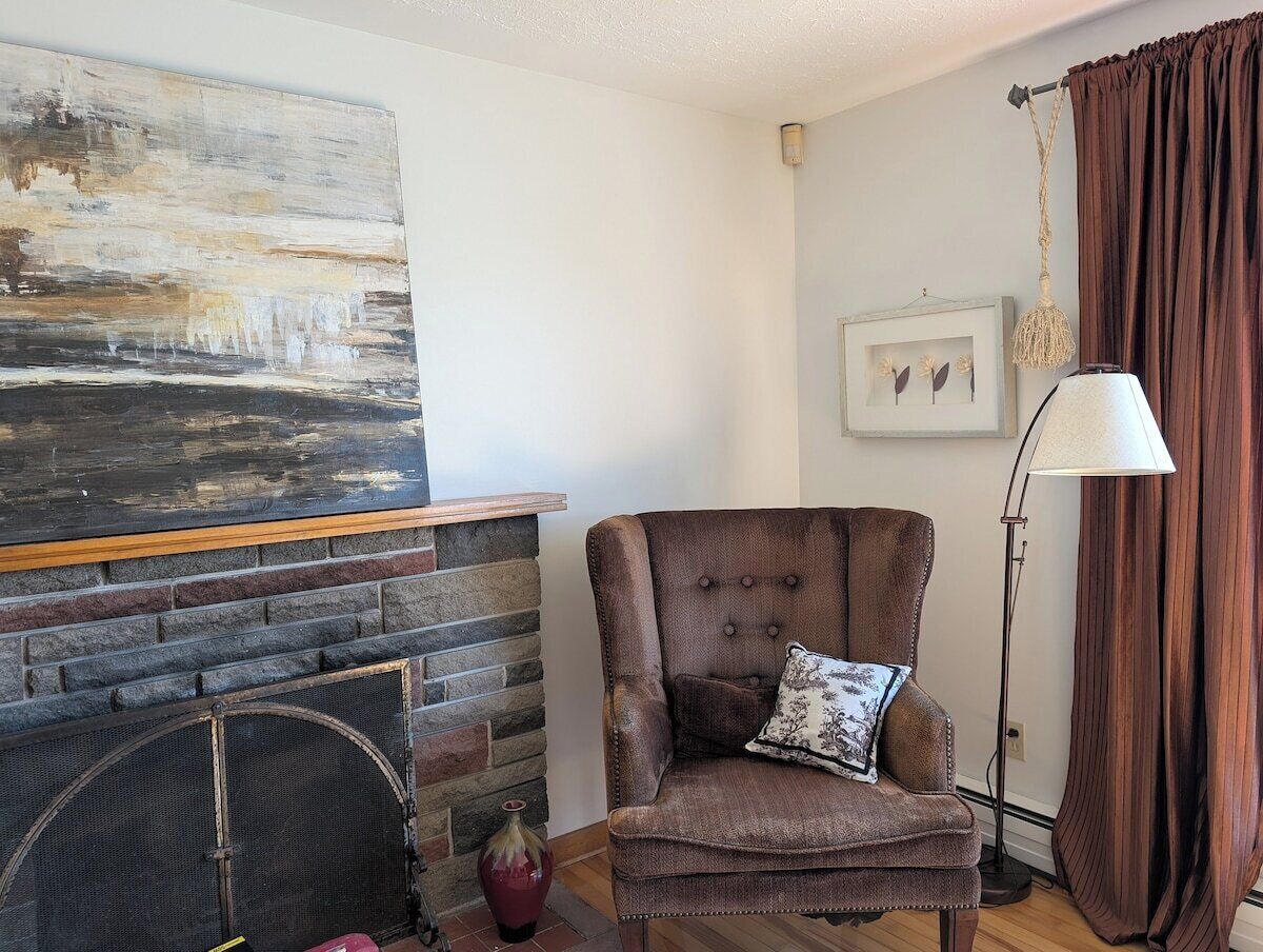 A living room with a fireplace, a brown armchair with a cushion, a floor lamp, and a large abstract painting on the wall.