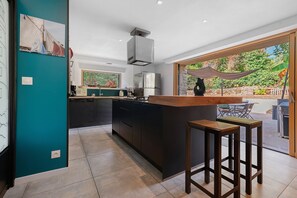 A modern kitchen with a central island, a dining area with a bar stool, and a view of the outdoors through a glass door. - Characterful House in the Gulf of Morbihan with Private Terrace and Garden (Sarzeau)