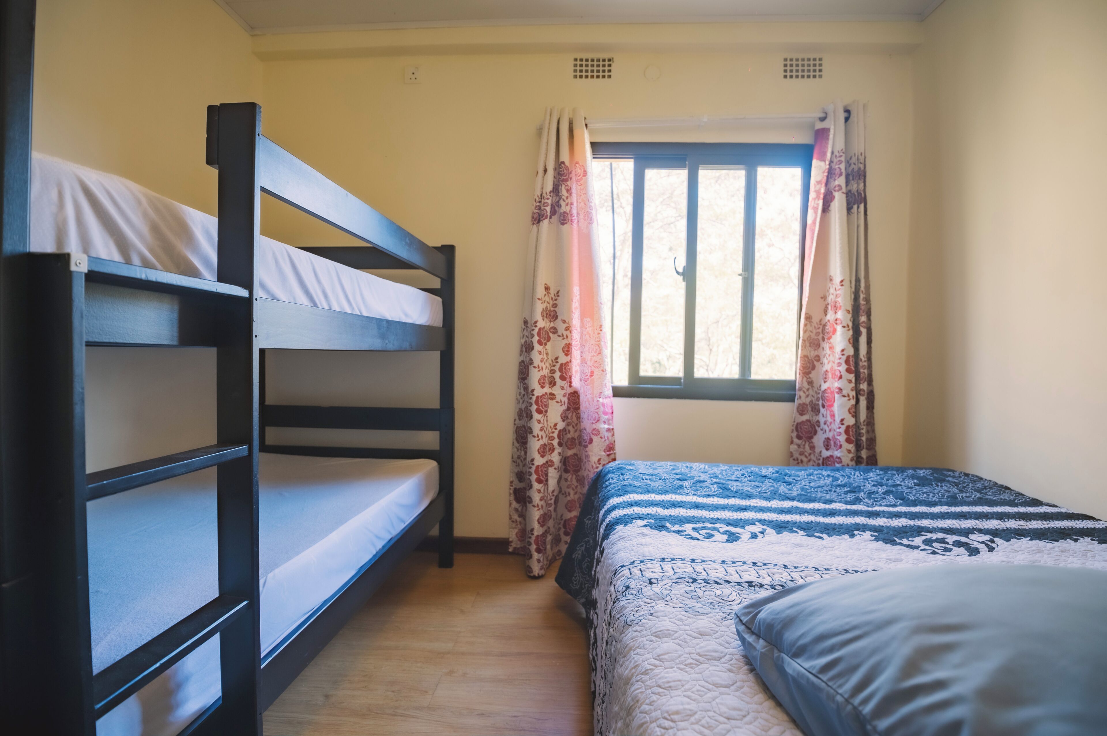 A room with bunk beds, a window with floral curtains, and a bed with a patterned bedspread.