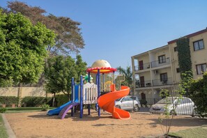 A playground with slides and climbing structures, surrounded by a sandy area and a building with balconies. - Beautiful 2-bedroom apartment in gated and secure community (Lusaka)