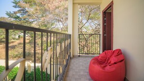 Uma varanda com uma poltrona de grãos vermelha, guarda-corpo de metal e vista para árvores e um edifício ao fundo.
