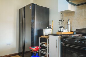 A compact kitchen with a refrigerator, a stove, a blender, and a small shelf with a basket. - Beautiful 2-bedroom apartment in gated and secure community (Lusaka)