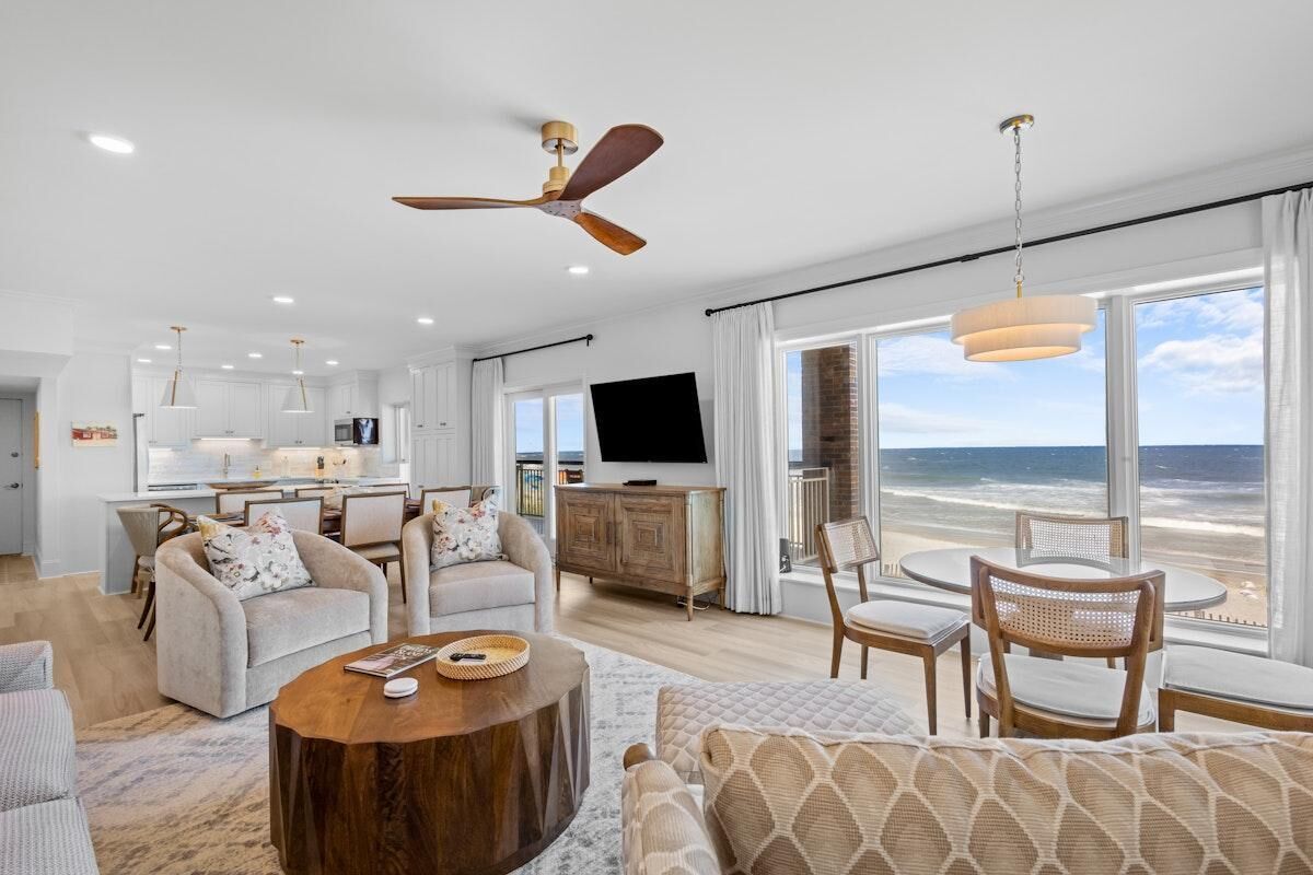 Een moderne woonkamer met een groot raam dat uitzicht biedt op het strand, een eethoek en een keuken met witte kasten.