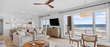 Een moderne woonkamer met een groot raam dat uitzicht biedt op het strand, een eethoek en een keuken met witte kasten.