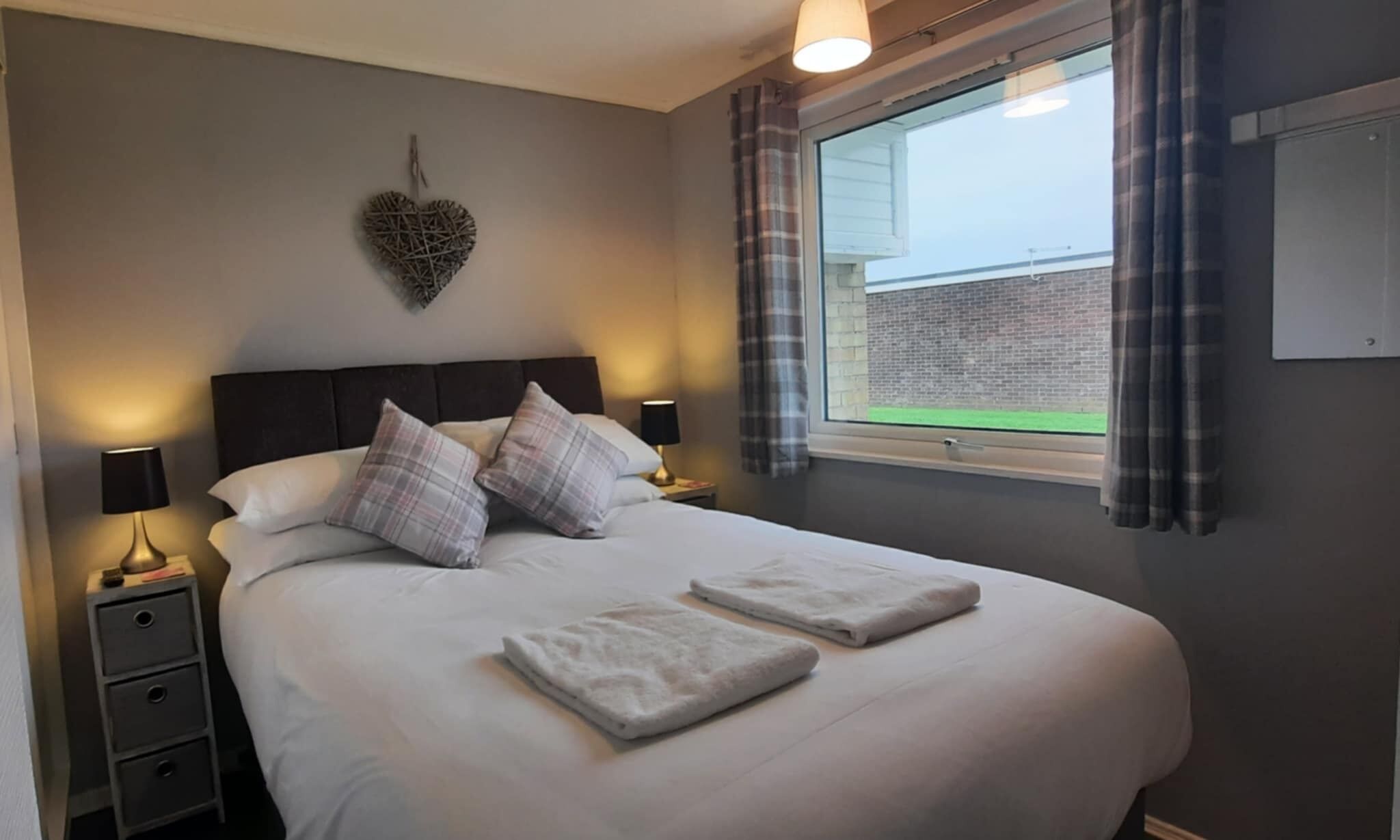 A neatly made bed with white linens and two pillows, a bedside lamp, a small bedside table, and a heart-shaped decoration on the wall.