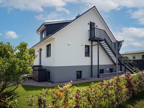 A modern two-story building with a flat roof, a balcony with a black railing, and a small outdoor area with grass and plants. - "Poeler Küstenkoje" Vacation Rental for 4 People (Insel Poel / OT Kirchdorf)