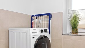 A compact, white front-loading washing machine with a Samsung label, positioned in a corner of a room with beige walls and a window. - "Poeler Küstenkoje" Vacation Rental for 4 People (Insel Poel / OT Kirchdorf)