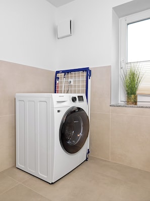 A compact, white front-loading washing machine with a Samsung label, positioned in a corner of a room with beige walls and a window. - "Poeler Küstenkoje" Vacation Rental for 4 People (Insel Poel / OT Kirchdorf)