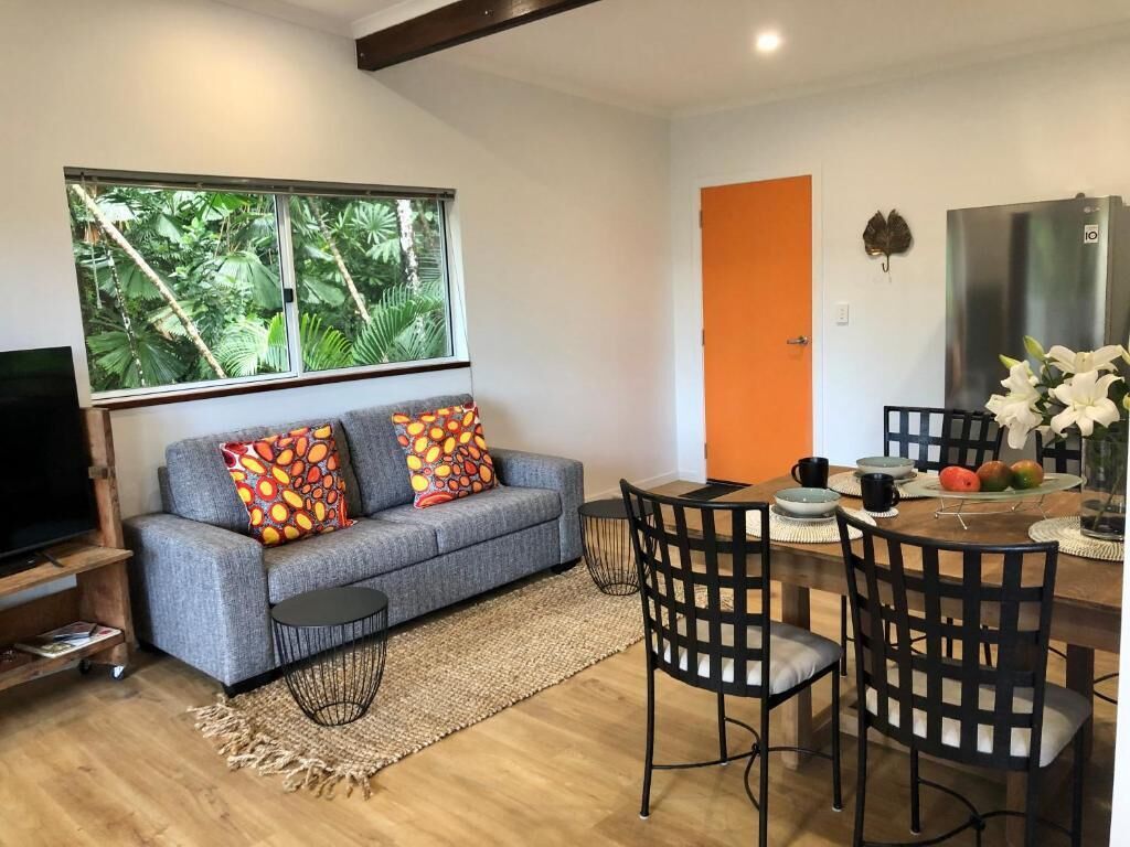 A modern living room with a grey sofa, a dining table with chairs, a large window with a view of greenery, and a kitchen area with a refrigerator.