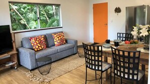 En moderne stue med en grå sofa, et spisebord med stole, et stort vindue med udsigt over grønne områder, og et køkken med et køleskab.