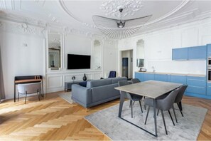 A modern living room with a sofa, dining table, and chairs, a television, and a kitchen area with blue cabinets. - Very spacious, recently renovated apartment near Madeleine (Île-de-France)