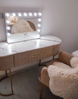 A vanity with a marble countertop, a fluffy chair, and a mirror with lights.