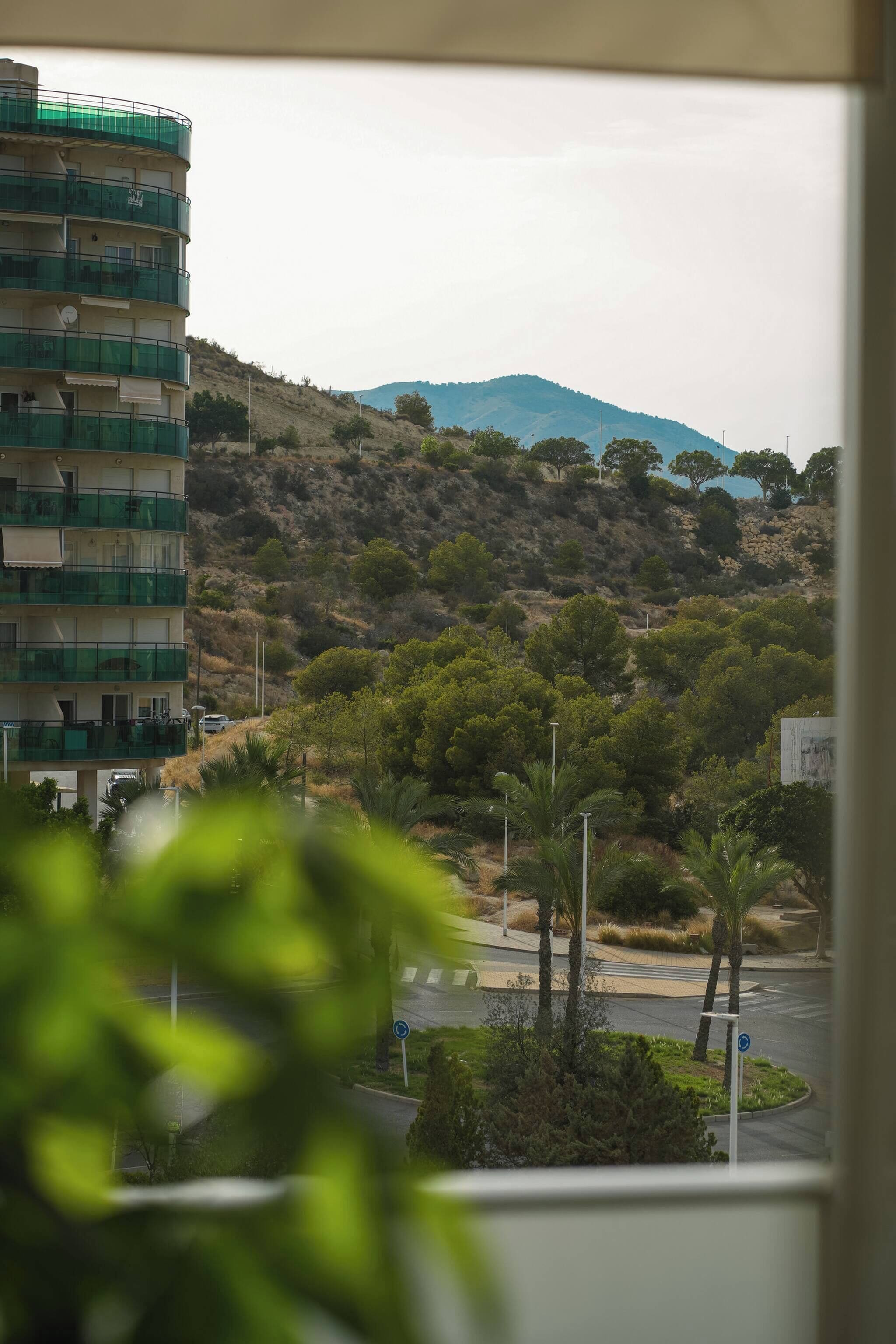Un edificio de varios pisos con balcones, un paisaje montañoso con árboles y una calle con paso de peatones.
