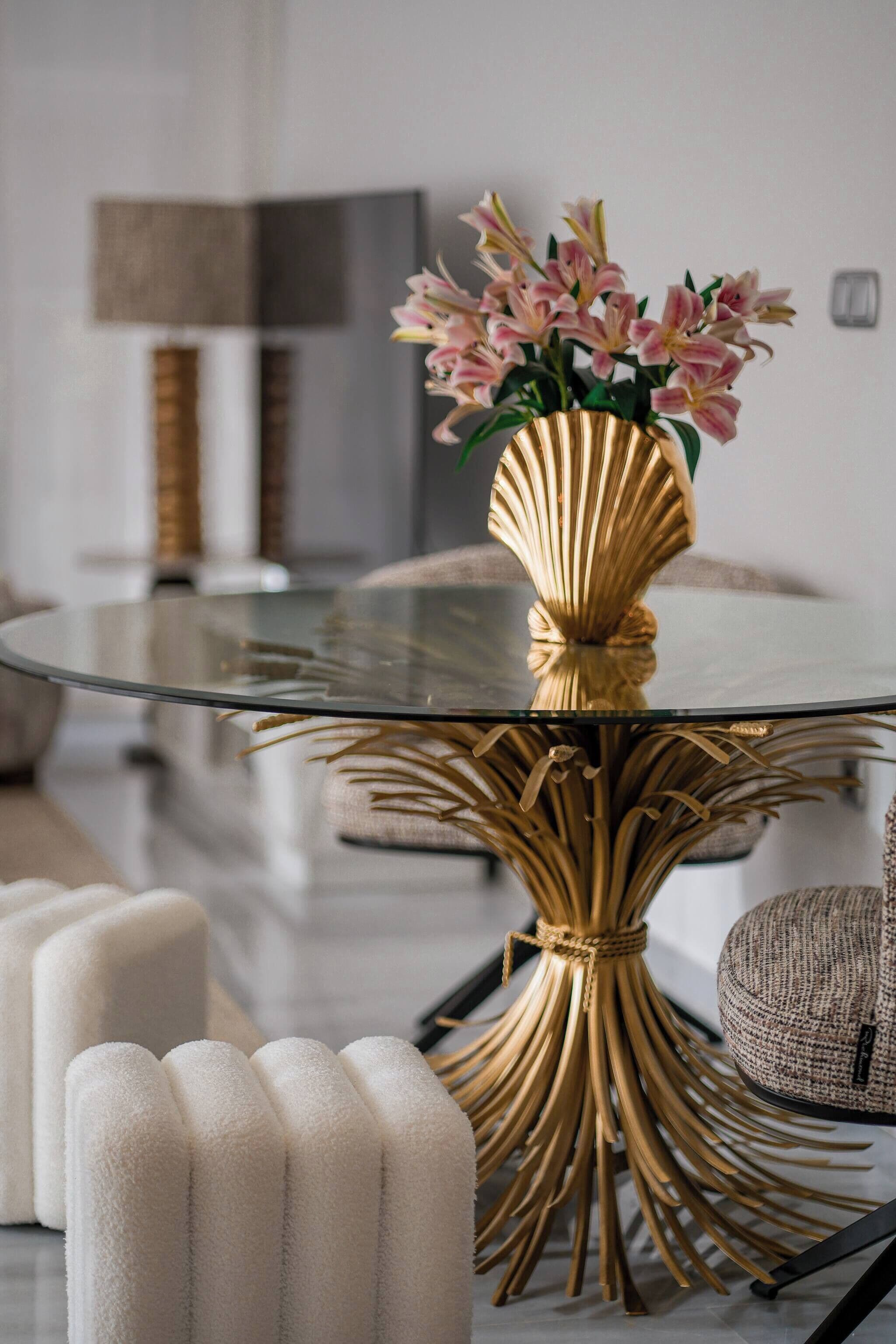 Ein runder Glastisch mit goldener Basis, umgeben von weißen, gepolsterten Poufs, und eine Vase mit rosa Blumen.