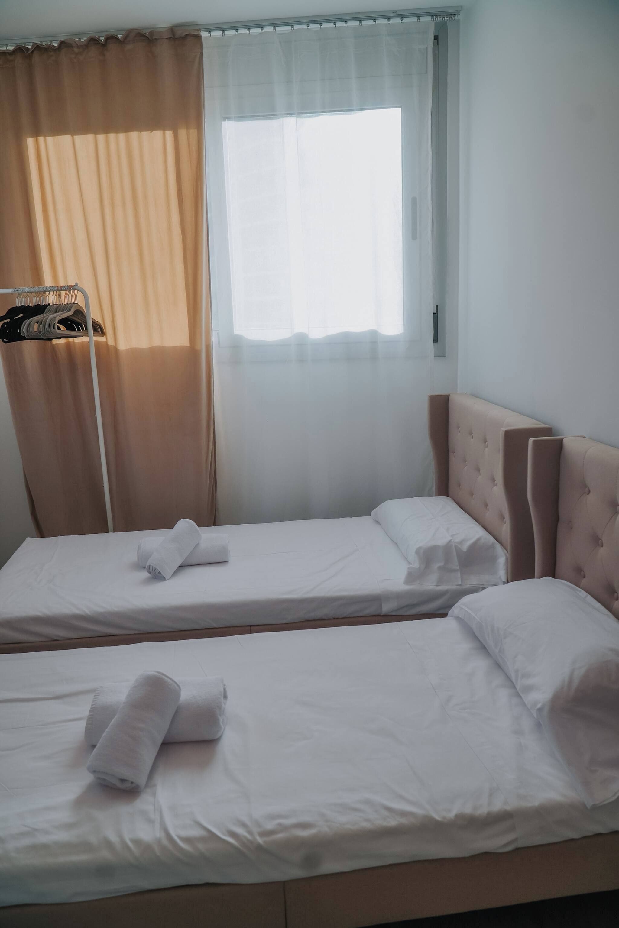 Habitación de hotel con dos camas individuales, una ventana con cortinas traslúcidas y un armario con ropa colgada.