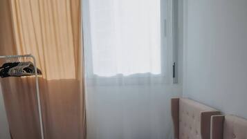 Habitación de hotel con dos camas individuales, una ventana con cortinas traslúcidas y un armario con ropa colgada.