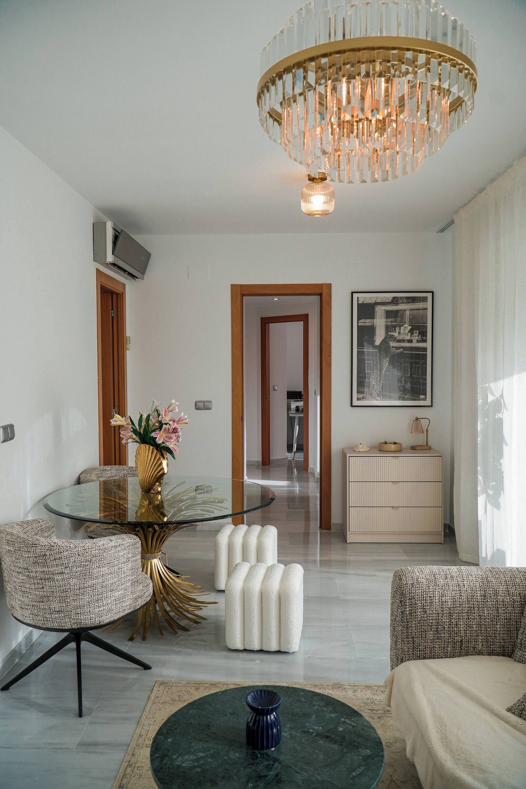 A modern living room with a glass coffee table, a round gold-framed table, and a white pouf.