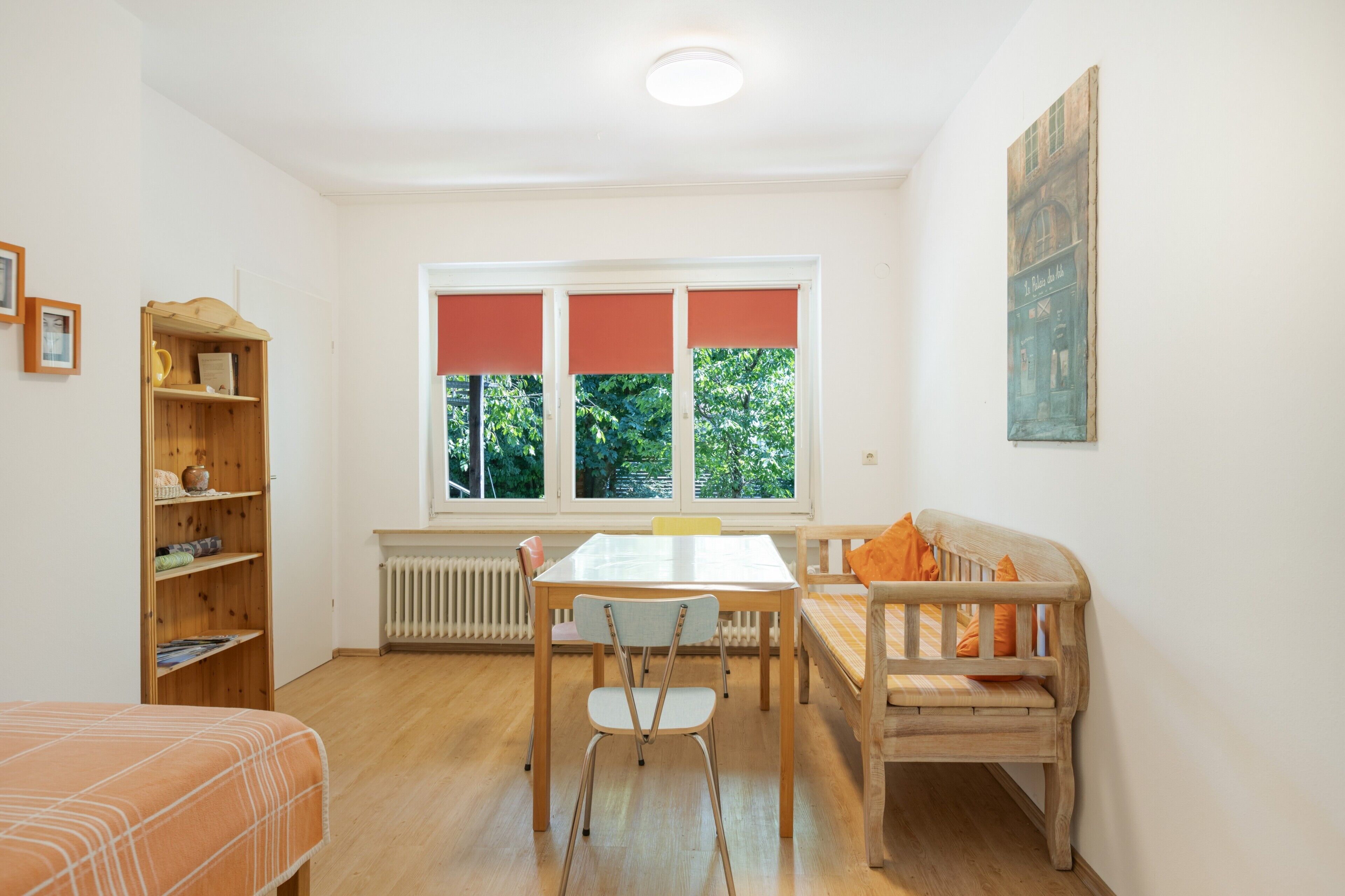 A room with a dining table, chairs, a sofa, a radiator, a wooden bookshelf, and a window with blinds.