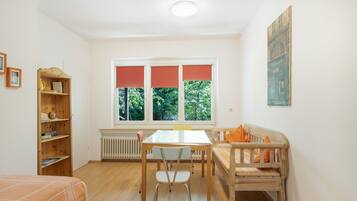 A room with a dining table, chairs, a sofa, a radiator, a wooden bookshelf, and a window with blinds.