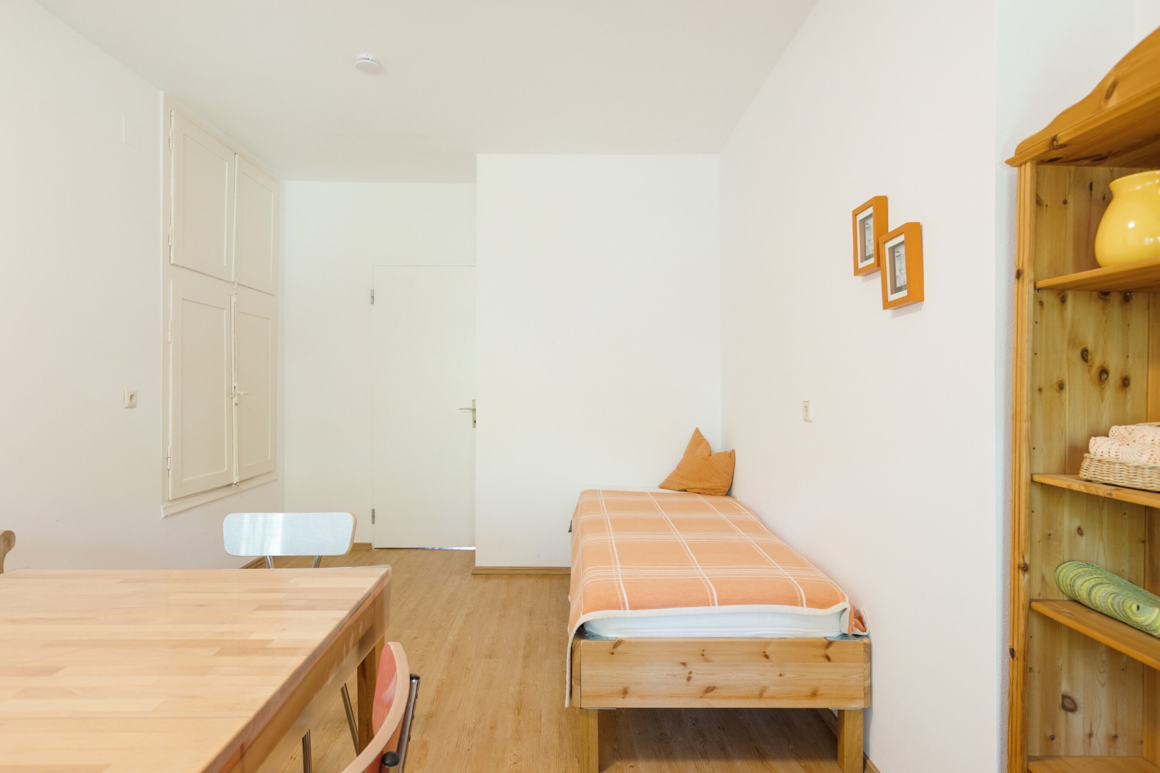 Ein kleiner, ordentlicher Raum mit einem Bett, einem Holztisch und einem Regal mit einem gelben Topf.