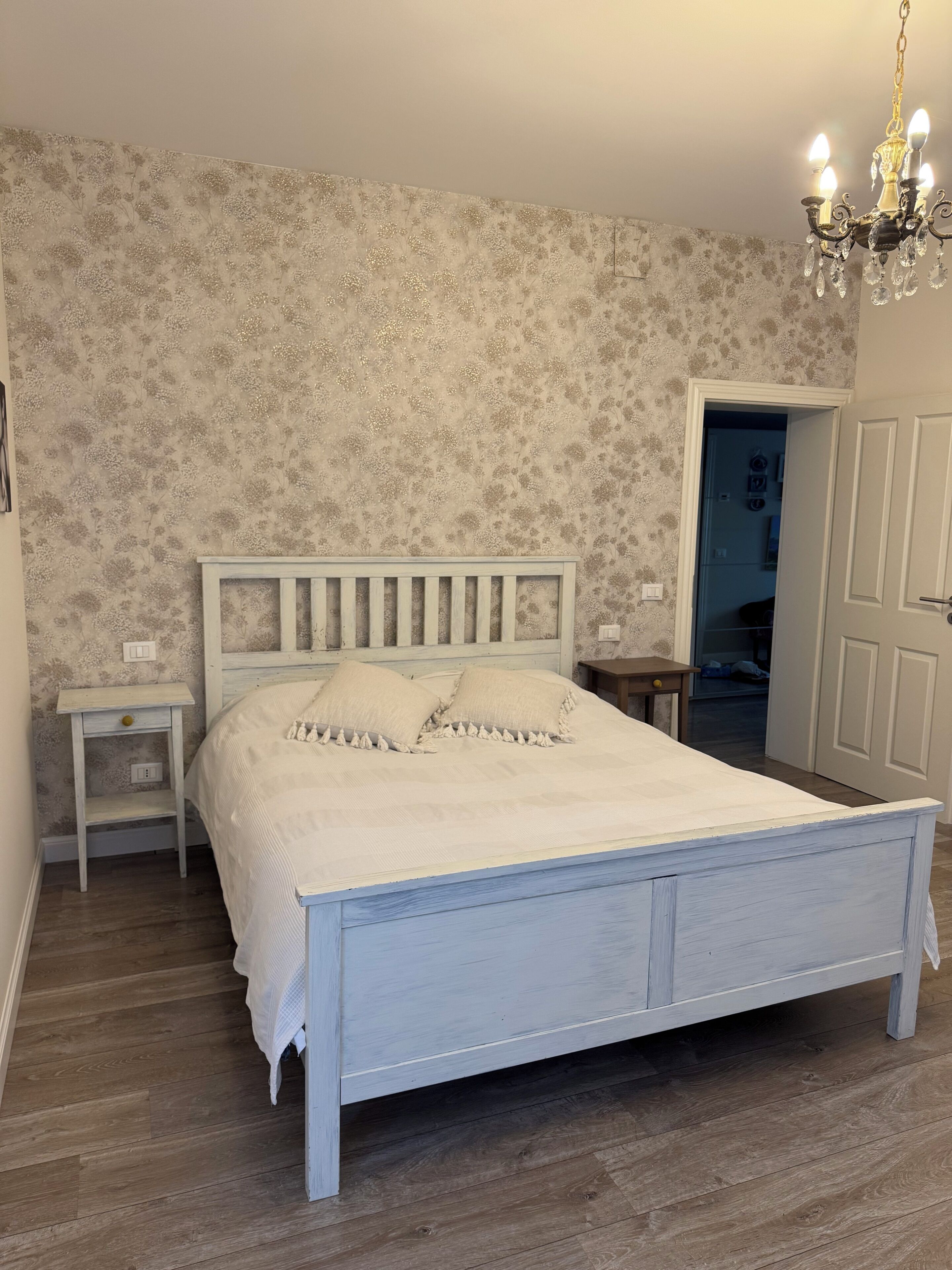 Ein Schlafzimmer mit Bett, Nachttisch, Schminktisch, einer Deckenleuchte und einer Tür zu einem anderen Raum.