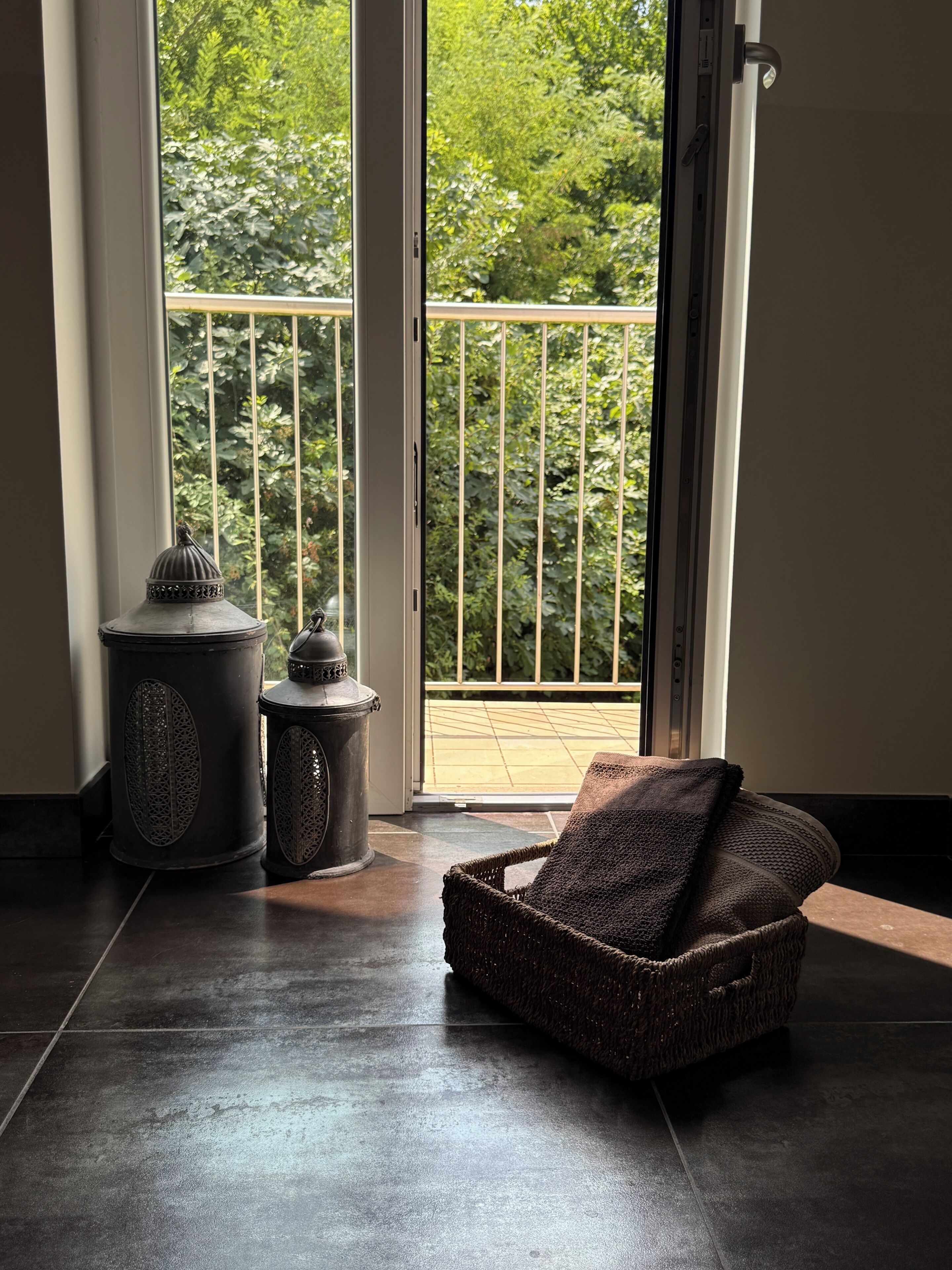 A room with a dark tiled floor, a wicker chair with a blanket, and two decorative metal containers near a sliding glass door with a view of greenery outside.