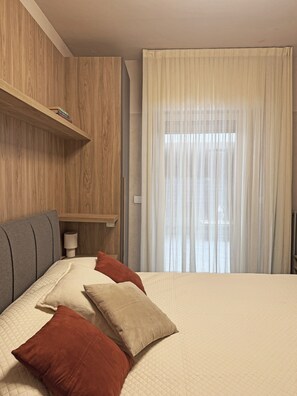 Ein Hotelzimmer mit einem Bett, Kissen, einem Nachttisch mit Lampe, einem Holzboden und einem Fenster mit Vorhängen.
