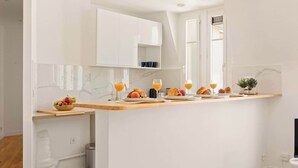 Une cuisine moderne avec des meubles blancs, un pare-feu en marbre et un coin repas où l’on sert des croissants, des fruits et du jus d’orange.
