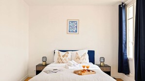 A neatly made bed with a blue headboard, two bedside tables with lamps, a framed artwork on the wall, and a window with dark curtains.