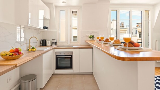 A modern kitchen with white cabinets, a built-in oven, a sink, and a breakfast setup with croissants and orange juice.