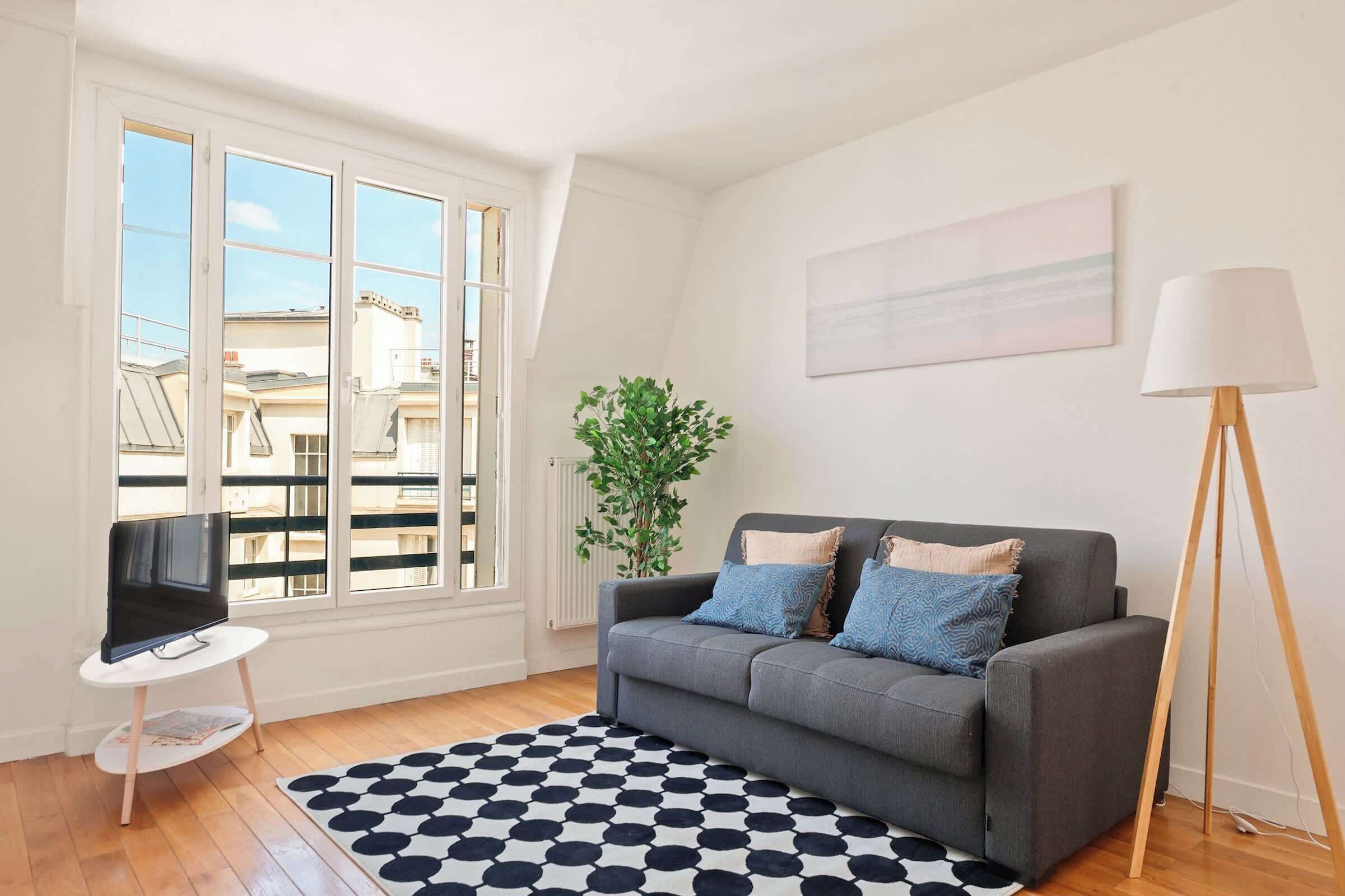 A living room with a grey sofa, a black and white patterned rug, a television on a white table, and a floor lamp.