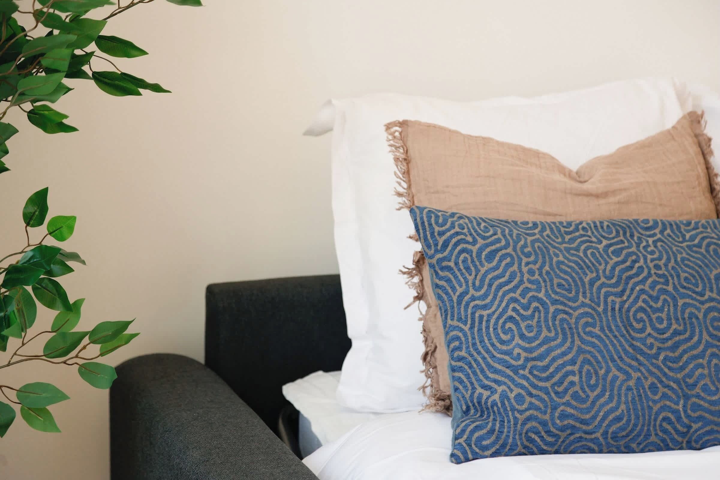 A bed with a white and beige blanket, a blue patterned pillow, and a dark-colored headboard.