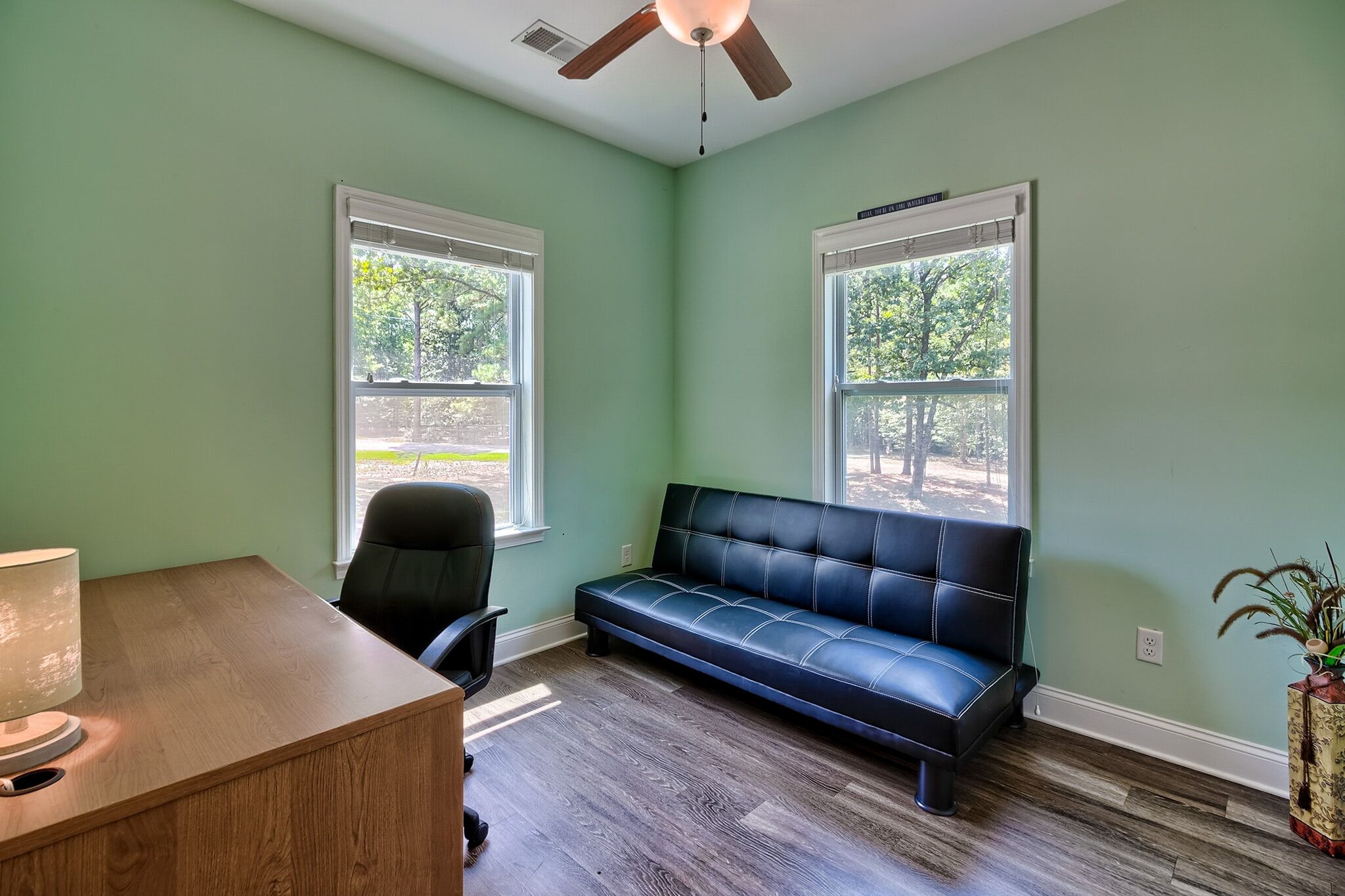 A room with a desk, a black leather sofa, two windows, and a ceiling fan.