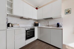 Cocina compacta con armarios blancos, horno empotrado, microondas y cafetera.