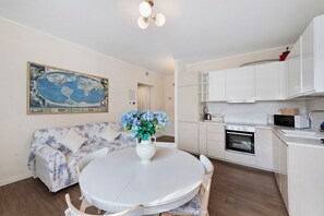 Una cocina moderna con gabinetes blancos, horno empotrado y microondas. Hay una mesa de comedor redonda con sillas y un florero con flores. Un mapa del mundo está colgado en la pared.