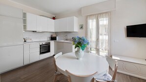 A modern kitchen with white cabinetry, a dining table with chairs, a vase with flowers, and a TV mounted on the wall. - ‘Elegant Lake Iseo’ Apartment with Lake & Mountain Views, Wi-Fi, Air Conditioning (Paratico)