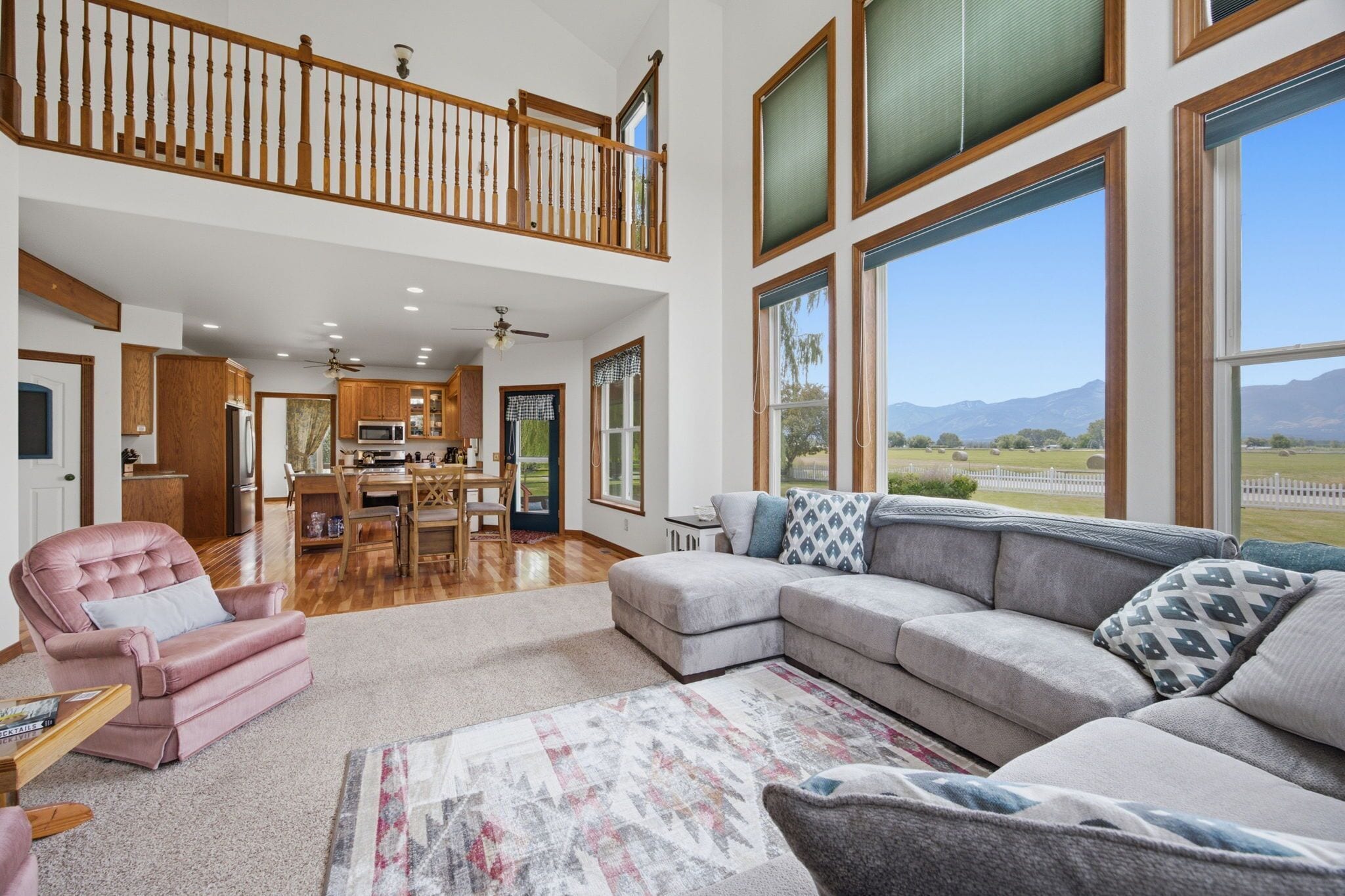 A spacious living room with a large sectional sofa, a pink armchair, and a dining area with wooden cabinets and a kitchen in the background.