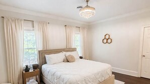 A bedroom with a large bed, a bedside table, a nightstand, a chandelier, and a window with curtains.