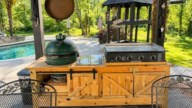 Estación de parrilla de madera al aire libre con parrilla, ahumador y chimenea, con vista a una piscina y abundante vegetación.