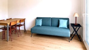 A room with a wooden dining table, two chairs, a blue sofa with cushions, a floor lamp, and a window. - Houlgate, near the beach, with outdoor space and parking (Houlgate)