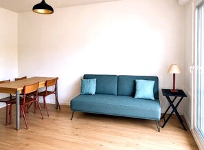 Ein Zimmer mit einem Holzesstisch, zwei Stühlen, einem blauen Sofa mit Kissen, einer Stehlampe und einem Fenster.
