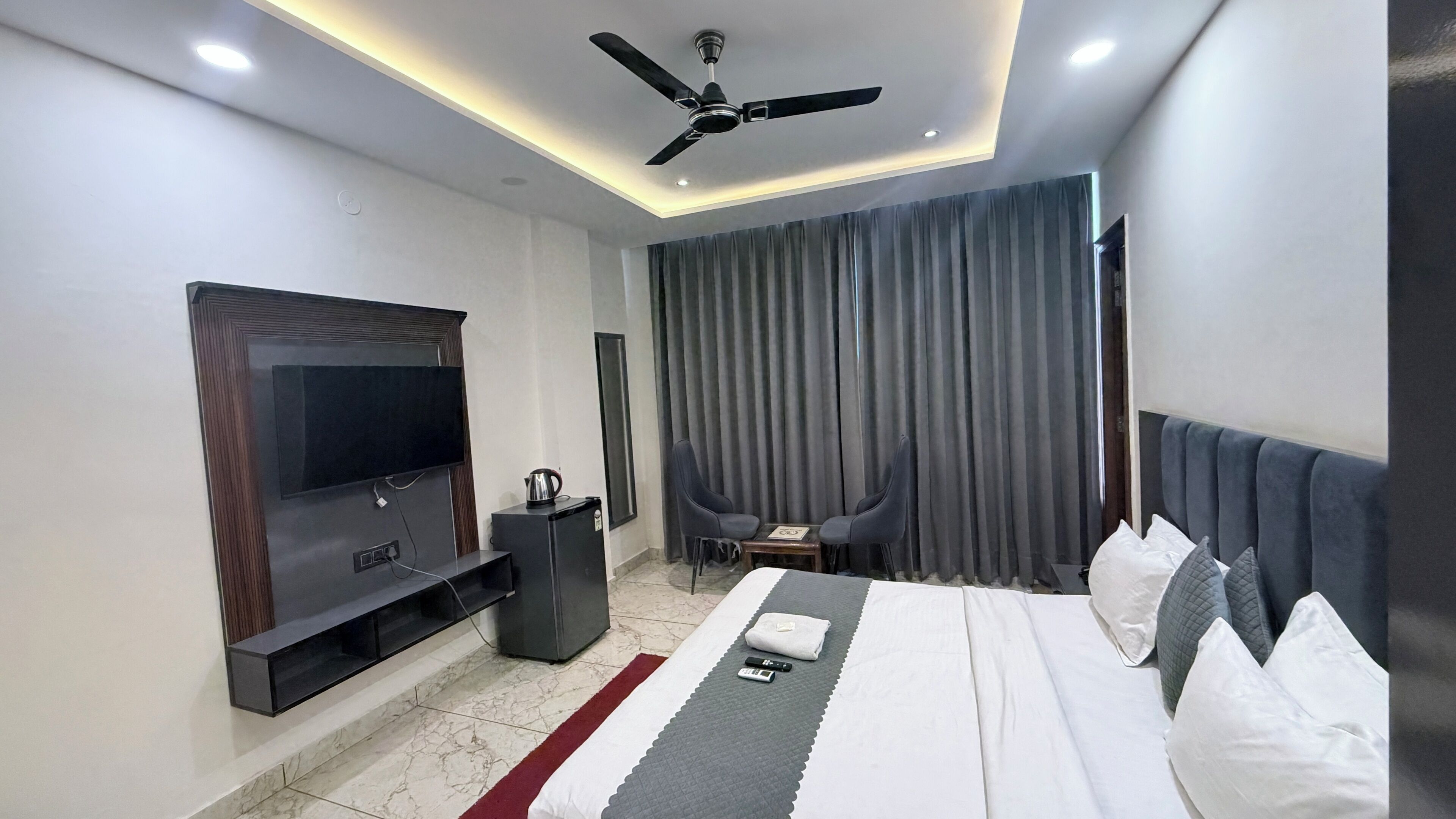 A modern hotel room with a flat-screen TV, a ceiling fan, a bed with pillows, a small table with a kettle, and a seating area with curtains.