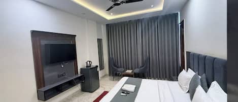 A modern hotel room with a flat-screen TV, a ceiling fan, a bed with pillows, a small table with a kettle, and a seating area with curtains.