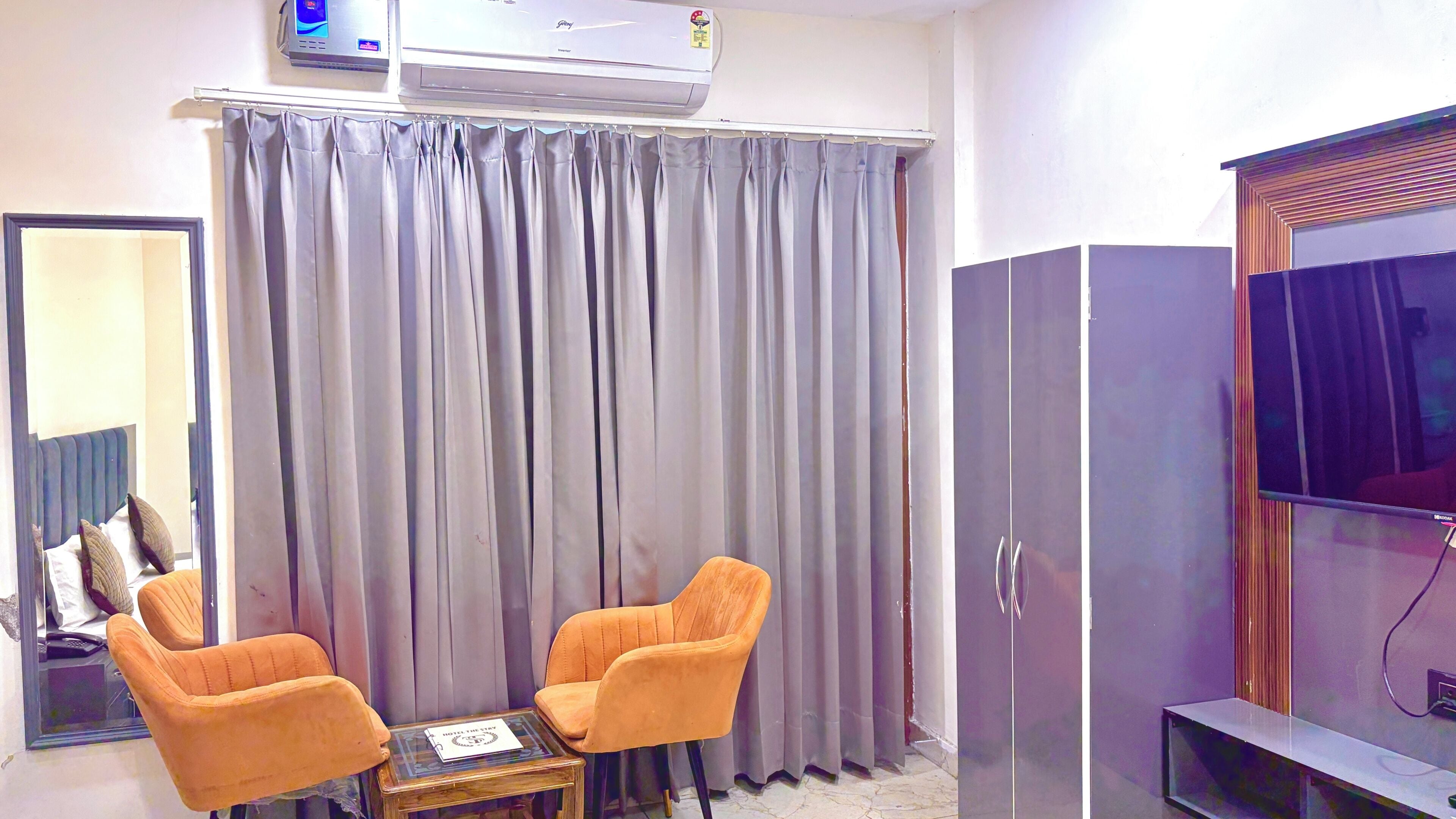 A room with a large grey curtain, two orange chairs, a small table with a book, a wardrobe, and a television.