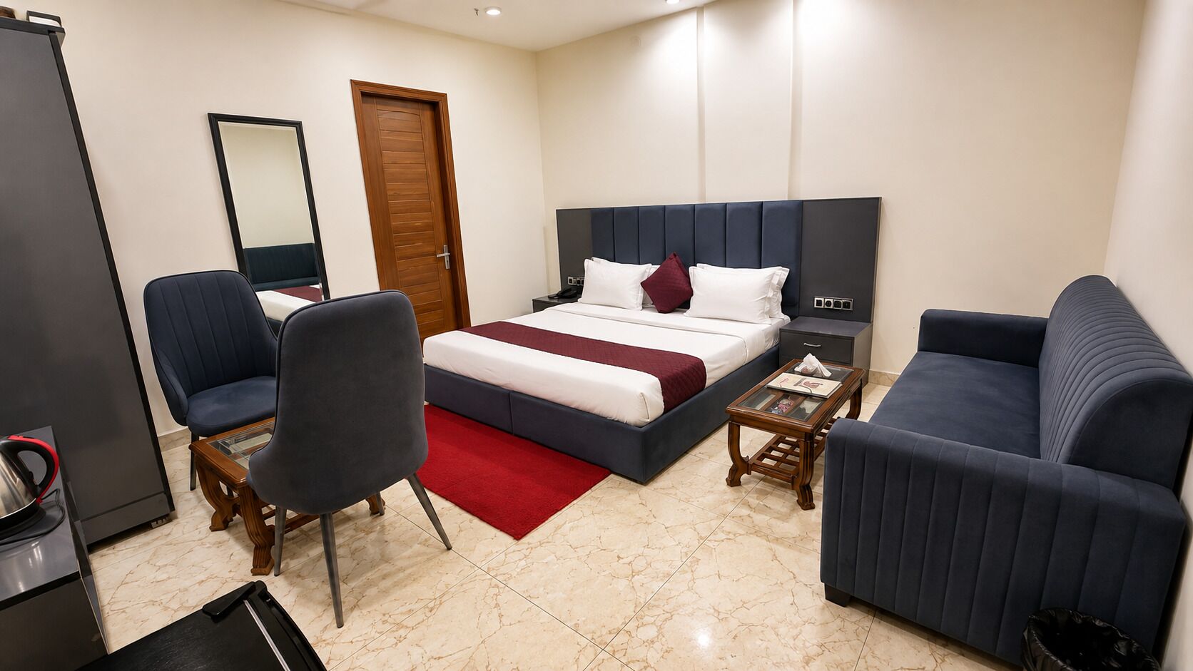 A hotel room with a bed, two armchairs, a small table, a nightstand, a mirror, and a door.