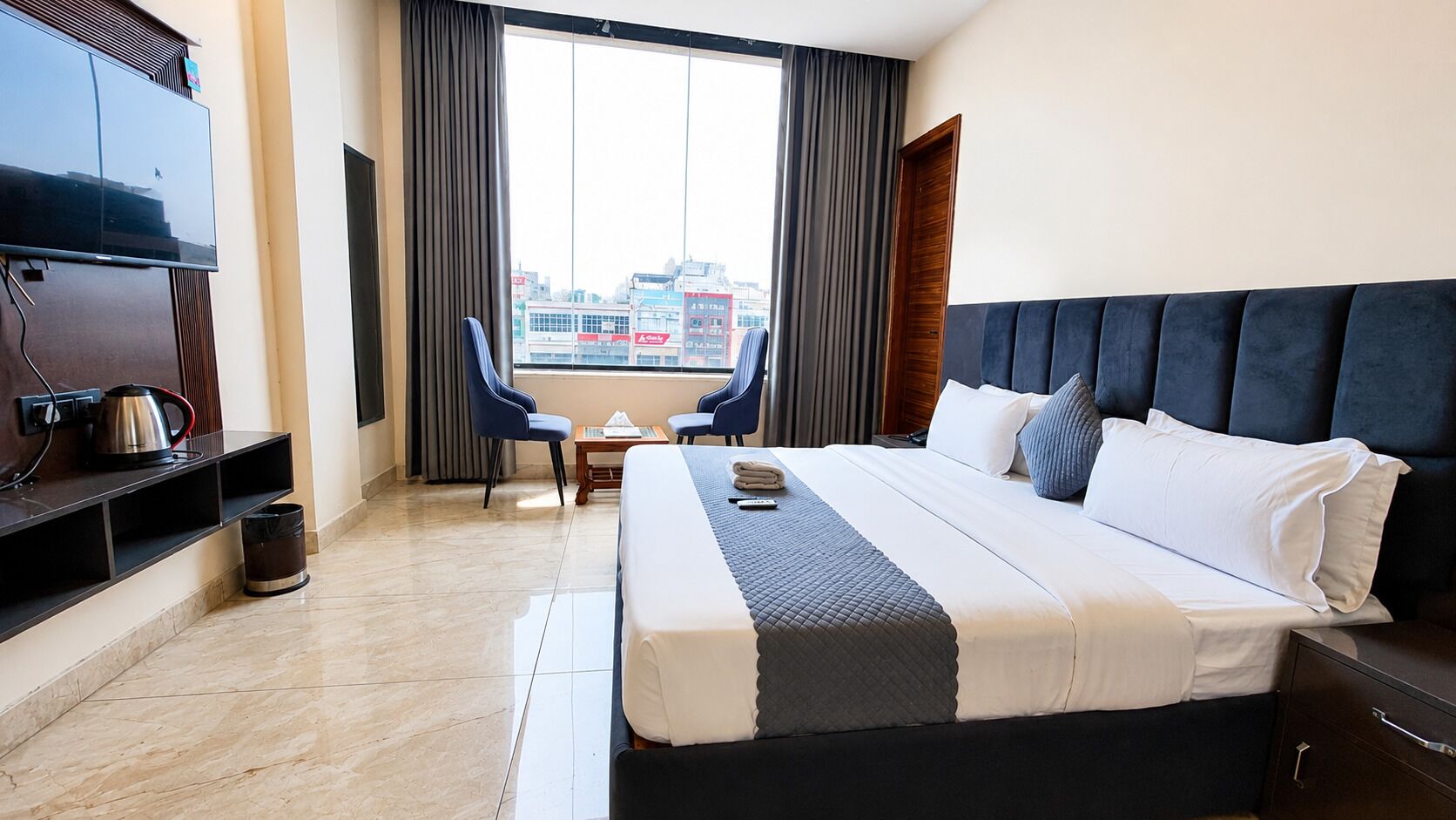 A hotel room with a bed, a TV, a coffee maker, a trash can, and a view of the city.