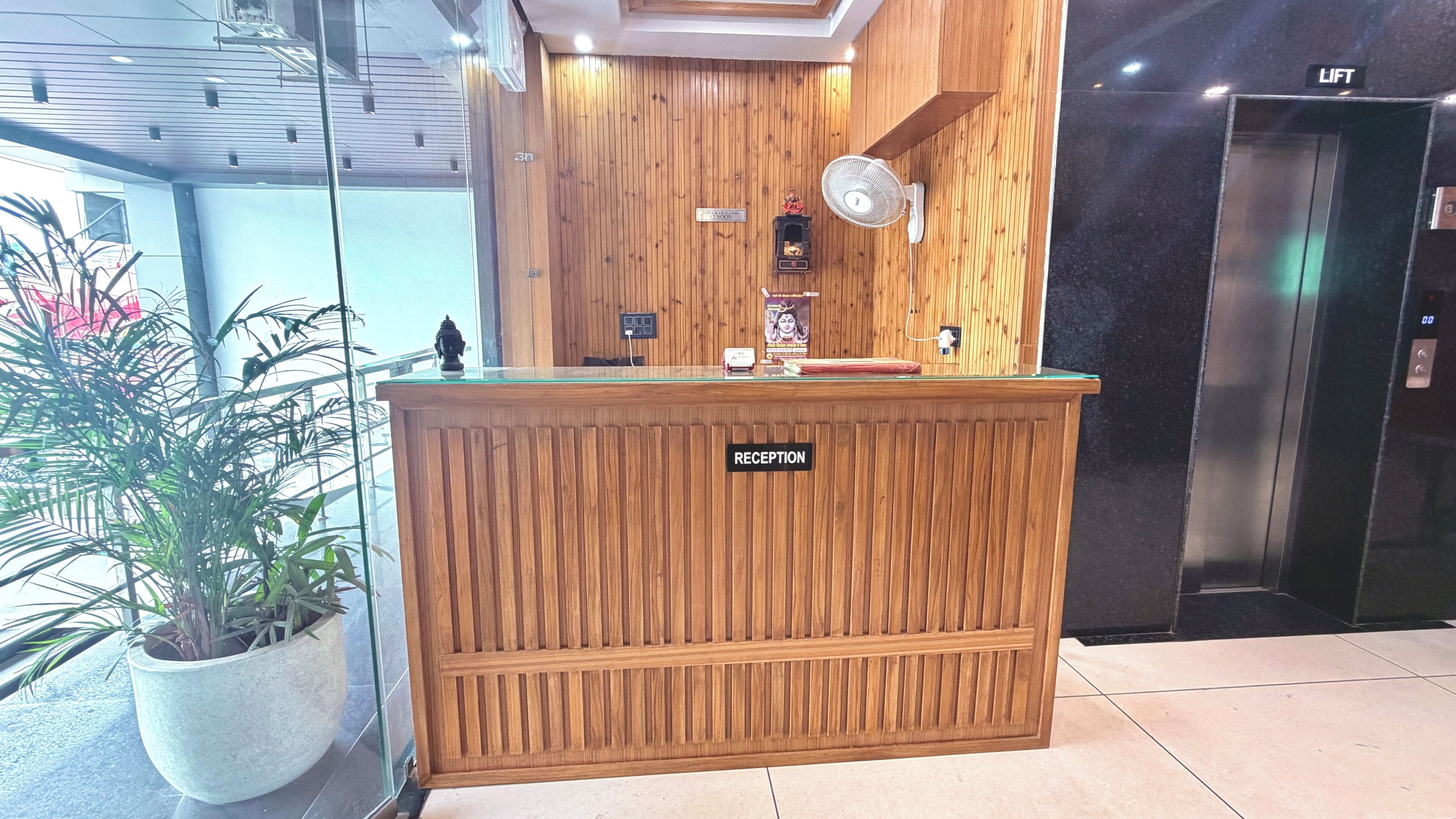 A reception desk with a 'RECEPTION' sign, a wooden wall, a fan, and a small statue.